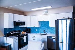 A modern kitchen with white cabinets, a stainless steel refrigerator, a microwave, a stove, and a sink with blue-tiled backsplash. - Recently Remodeled Beachfront Escape at Florence 1 #307 - Corner Unit (South Padre Island)