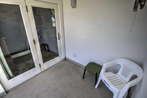 Una habitación con puerta de vidrio corredera que da a un balcón, un taburete pequeño verde y una silla de plástico blanca.