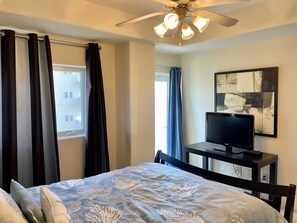 Une chambre à coucher avec un lit, un téléviseur posé sur un meuble, un ventilateur de plafond et une fenêtre avec des rideaux.