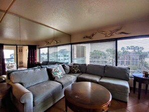 A living room with a sectional sofa, a round coffee table, and a view through large windows with blinds. - Landfall Towers #34 - Stunning Bayfront Living with Private Boat Slip Access (South Padre Island)