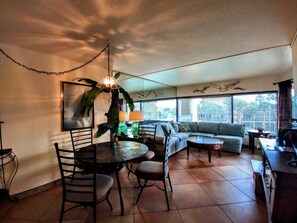 A living room with a dining table, chairs, a sofa, and a coffee table. There is a large window with blinds, a hanging plant, and a wall-mounted lamp. - Landfall Towers #34 - Stunning Bayfront Living with Private Boat Slip Access (South Padre Island)