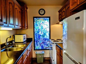 A compact kitchen with wooden cabinets, a stainless steel sink, a microwave, a stove, and a refrigerator. A clock is mounted on the wall above a stained glass window. - Landfall Towers #34 - Stunning Bayfront Living with Private Boat Slip Access (South Padre Island)