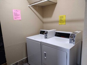 A laundry area with two front-loading washing machines, a pink sign listing laundry items and prices, and a yellow sign advising not to wash sandy clothes. - Landfall Towers #71 - Breathtaking Bayfront Living with Boat Slip Access (South Padre Island)
