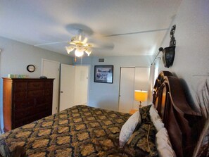 A bedroom with a patterned bedspread, a wooden dresser, a ceiling fan, a wall-mounted lamp, and a framed picture. - Landfall Towers #71 - Breathtaking Bayfront Living with Boat Slip Access (South Padre Island)