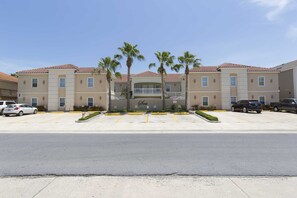 Een meerlaags hotel met een parkeerplaats, palmbomen en een heldere lucht.