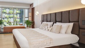 Quarto moderno com uma cama grande, um banco de madeira e vista para o verde lá fora.