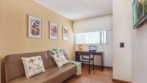 Sala de estar com sofá de couro marrom, duas almofadas decorativas, uma mesinha com um laptop e uma janela com vista.