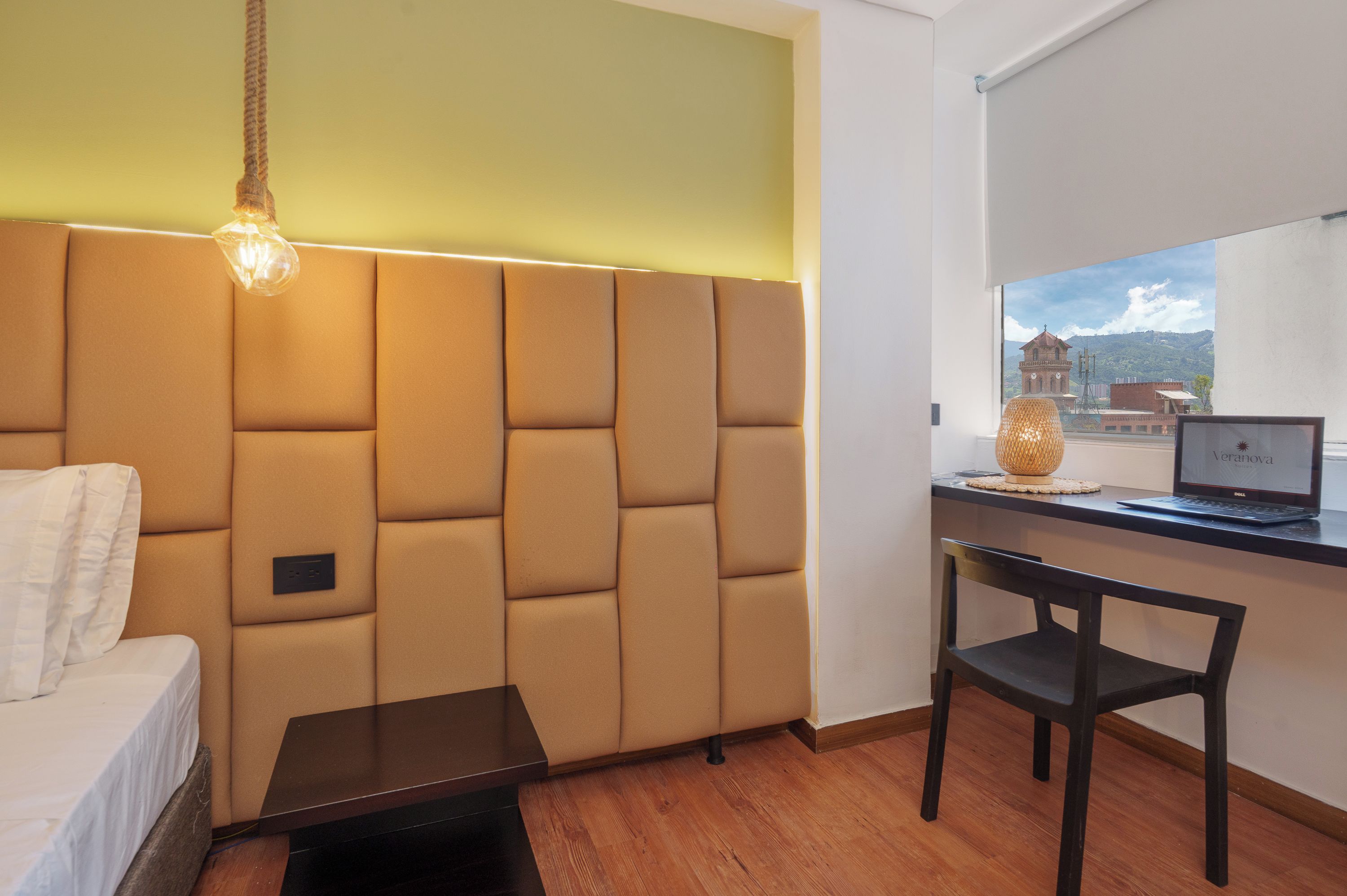 A room with a sofa, a desk, a laptop, and a view of a building through a window.