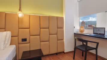 A room with a sofa, a desk, a laptop, and a view of a building through a window.