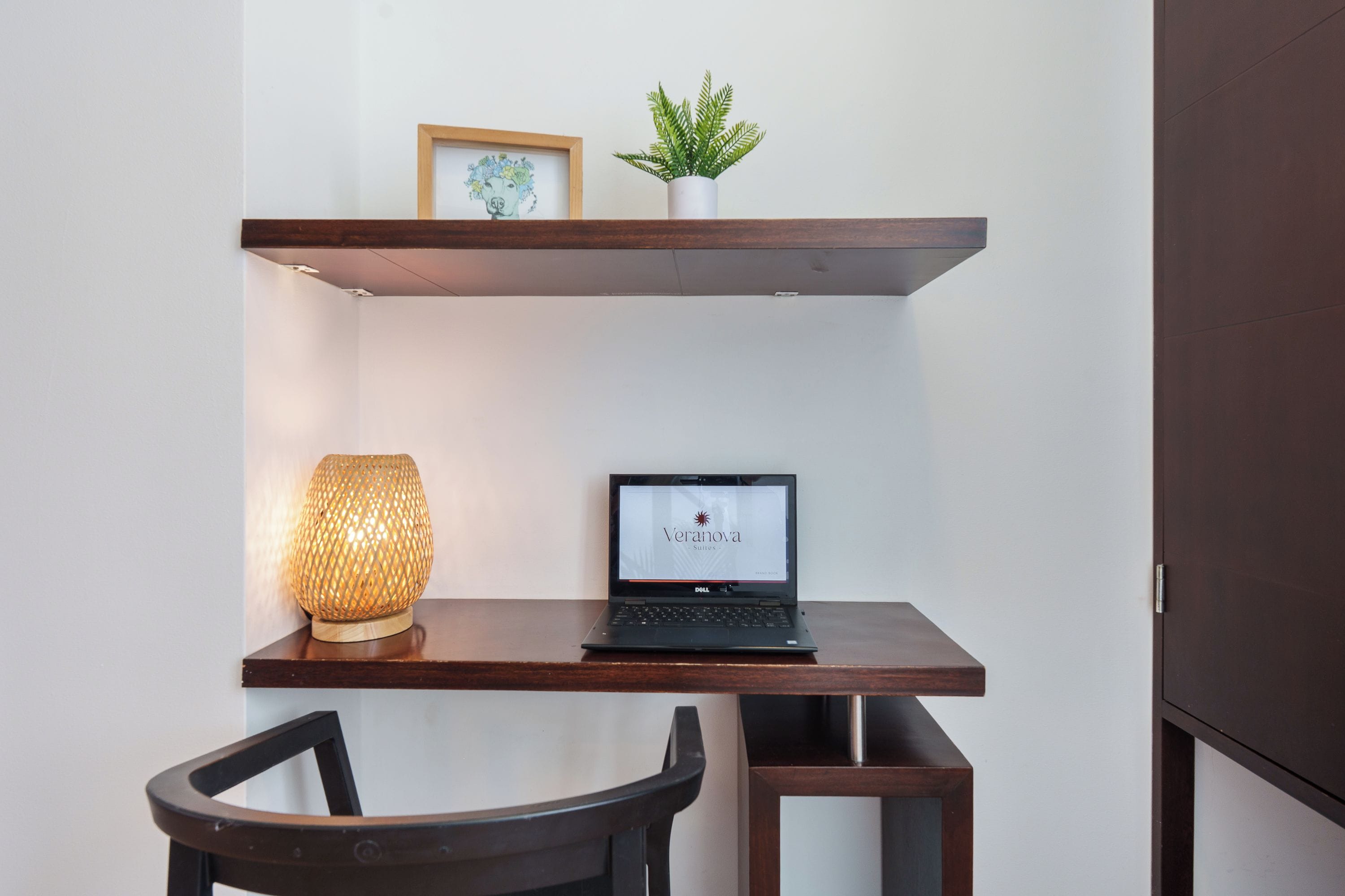 Un ordinateur portable est posé sur un bureau en bois, avec une lampe et une plante en pot sur une étagère située au-dessus.