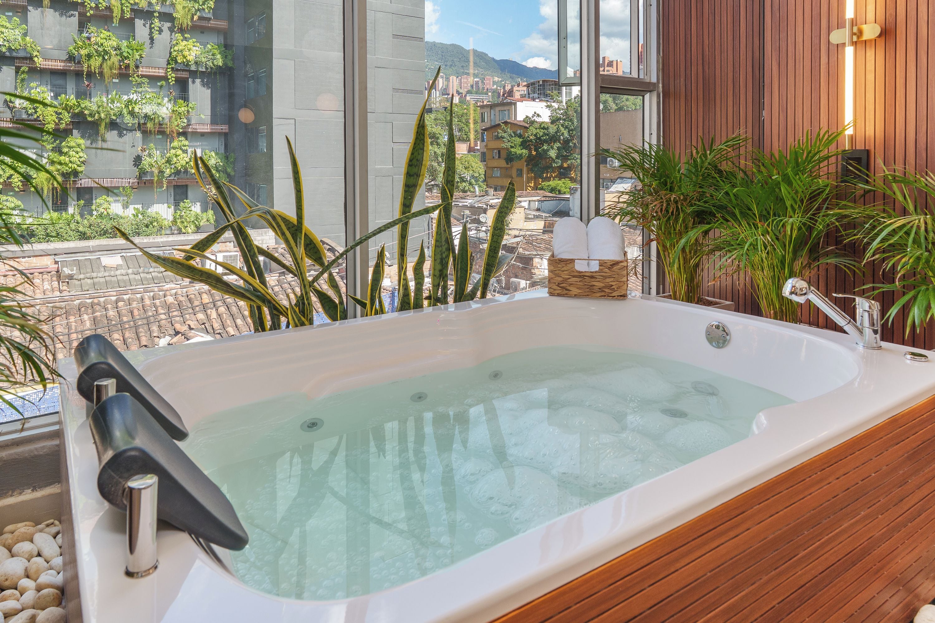 A hot tub with a wooden interior, a window with a view of buildings and greenery, and a small basket with towels.
