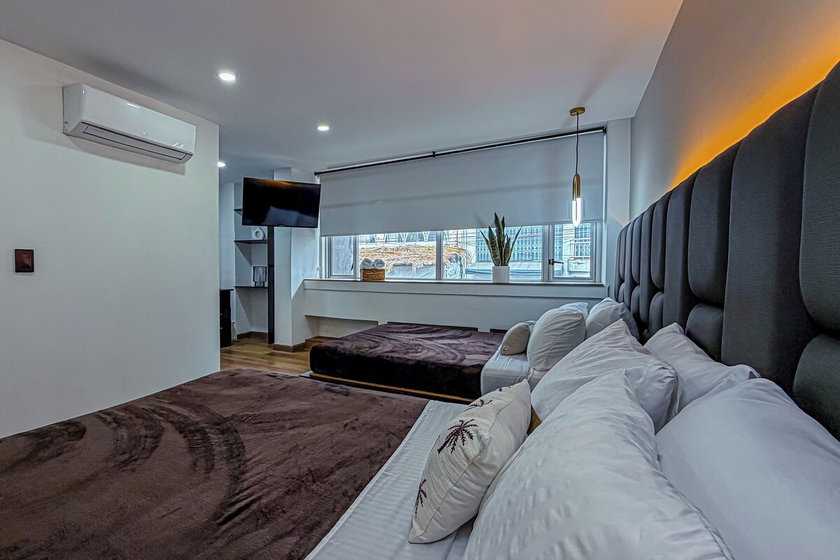 Un dormitorio moderno con una cama grande, una cama más pequeña, un televisor y una ventana con vistas a la ciudad.