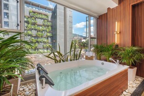 Un baño moderno con una bañera grande, paneles de madera y vistas de un paisaje urbano a través de amplios ventanales.