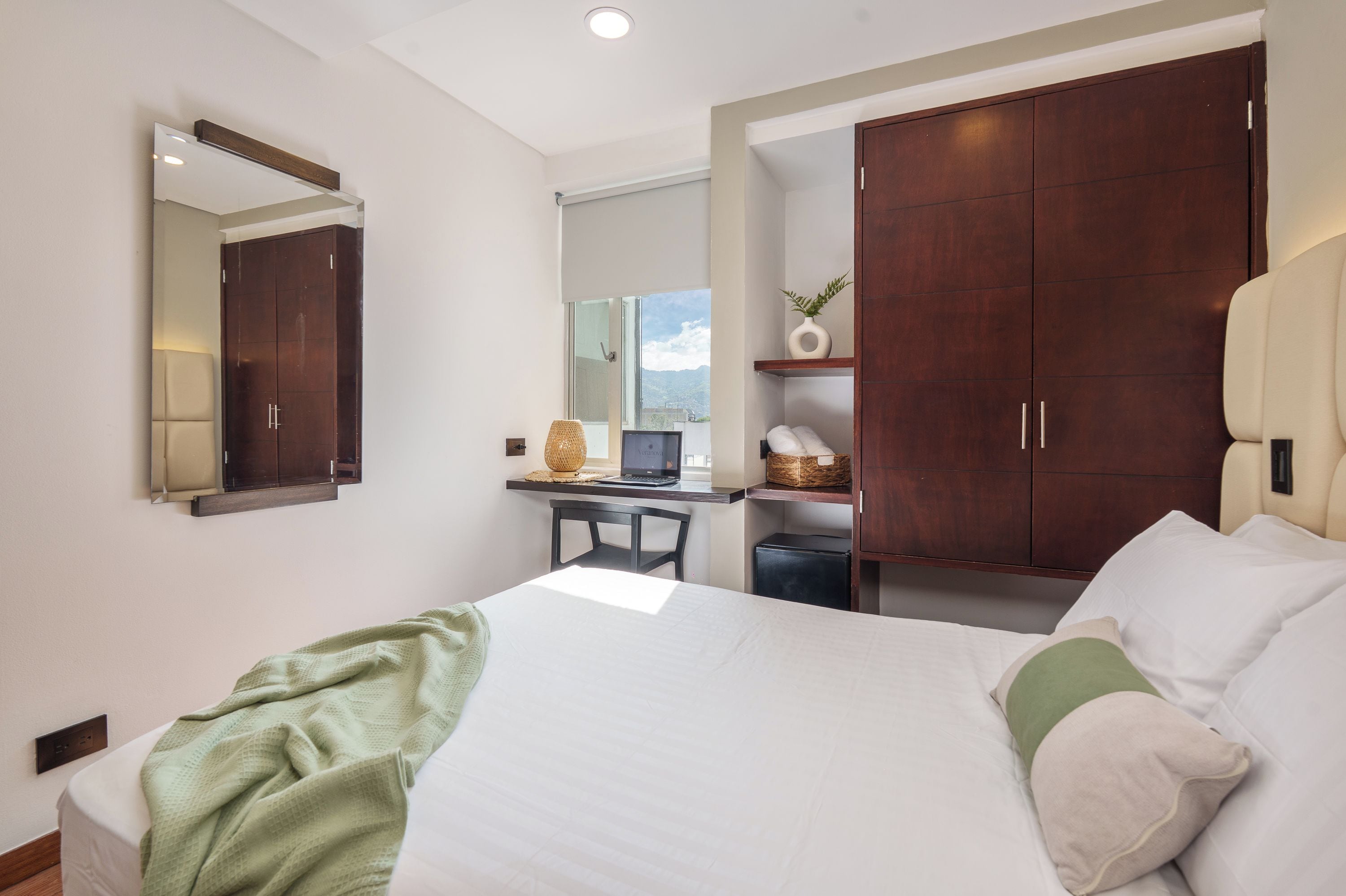Una habitación de hotel con una cama, un escritorio con ordenador, un armario y un espejo.