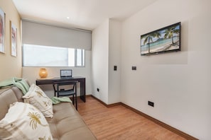 A modern living room with a sofa, a desk with a laptop, a painting of a beach scene, and a window with blinds. - Veranova hotel (Medellín)