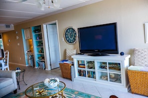 A living room with a television, a bookshelf, a wicker chair, and a patterned rug. - Seville #602 - Premier Top-Floor Beachfront Living with Panoramic Gulf Views (South Padre Island)