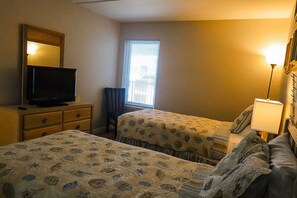 Ein Hotelzimmer mit zwei Betten, einem Fernseher auf einer Kommode, einem Fenster mit Jalousien und einer Lampe.