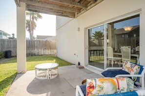 Un patio cubierto con una mesa redonda y sillas, una puerta corrediza que da a un área de comedor al aire libre, y una valla de madera al fondo.
