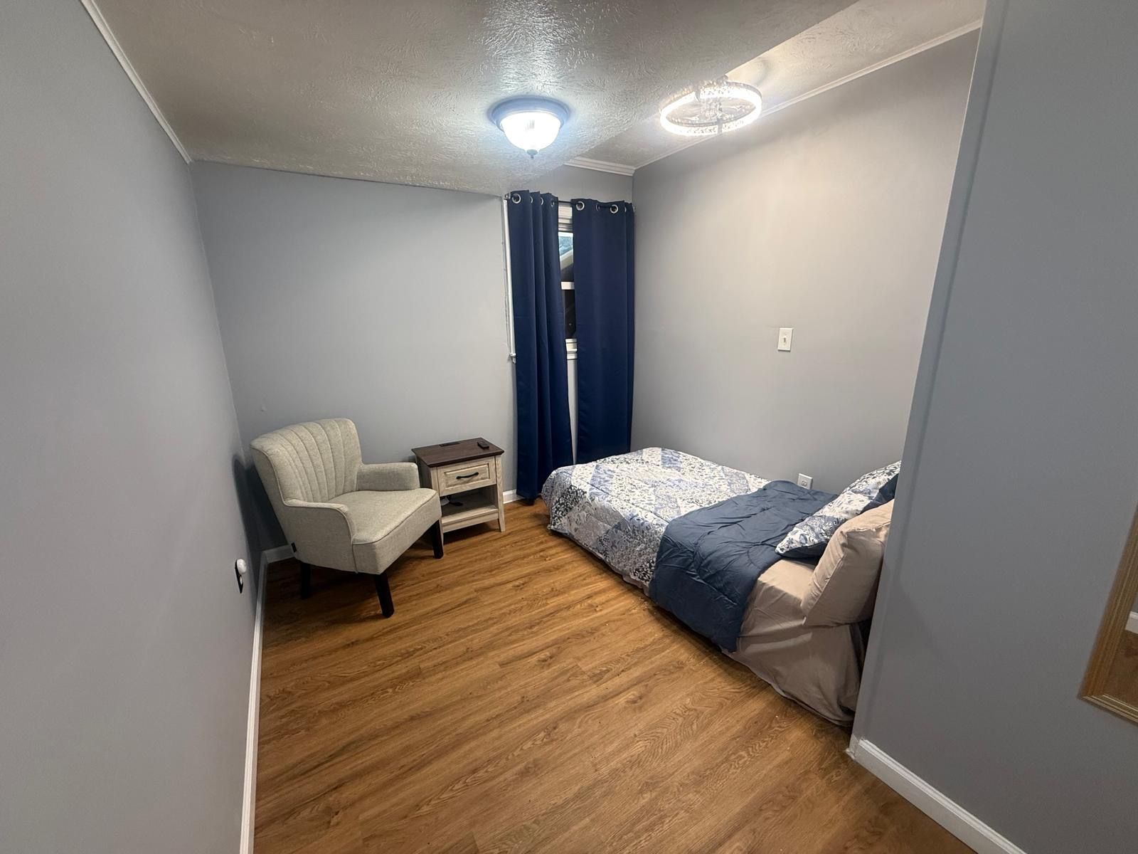 A small bedroom with a single bed, a chair, a nightstand, and a ceiling light fixture.