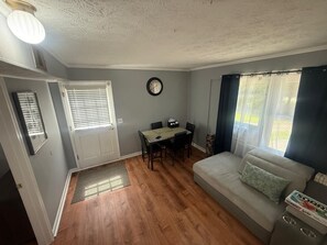 Una sala de estar con sofá, una mesa de comedor con sillas, un reloj en la pared y un ventanal con cortinas.