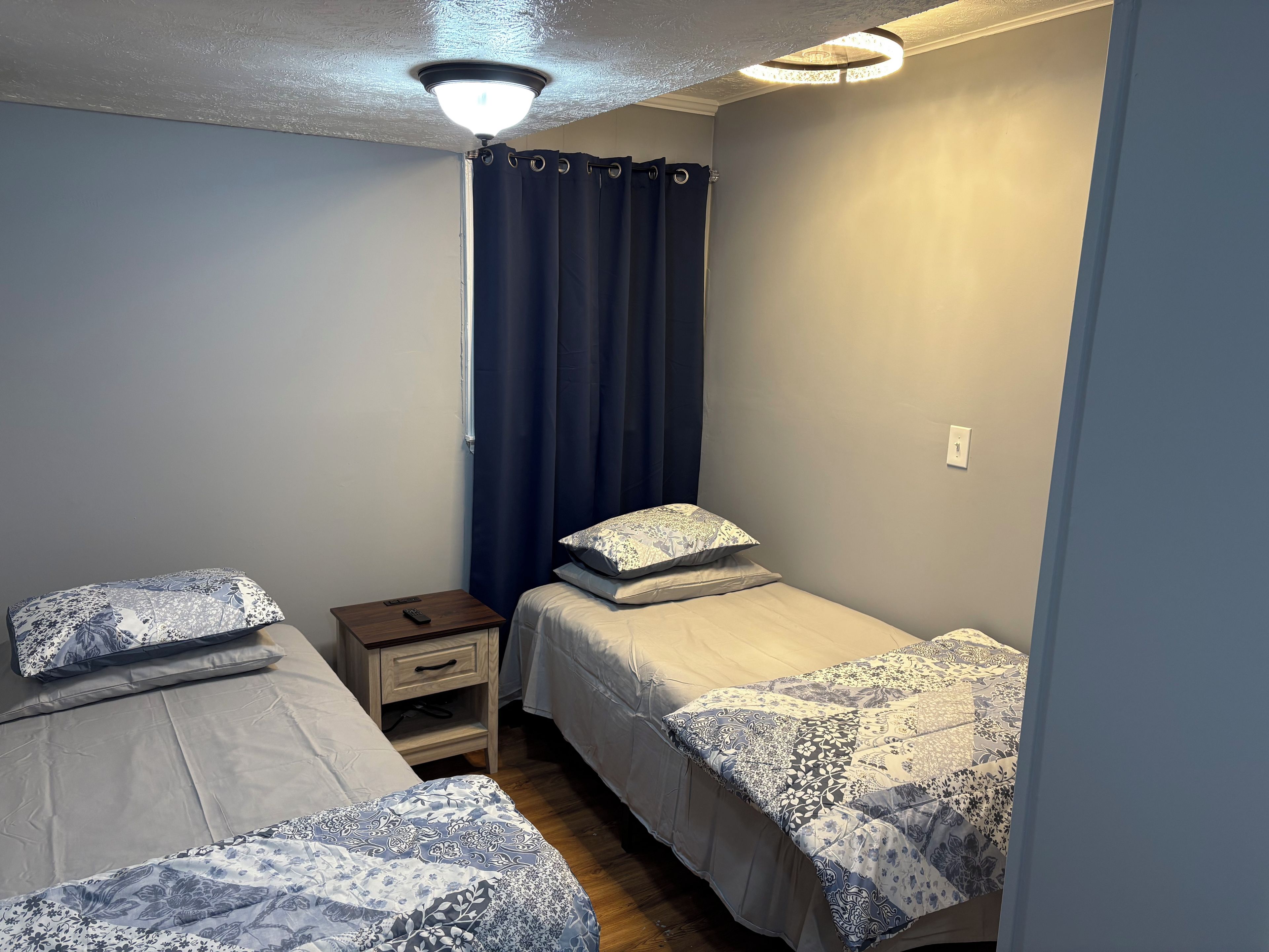 A small, well-lit bedroom with two beds, a nightstand, a ceiling fan, and a window with curtains.