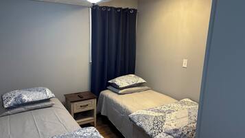 A small, well-lit bedroom with two beds, a nightstand, a ceiling fan, and a window with curtains.