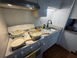 Una cocina con armarios blancos, un lavamanos de acero inoxidable y una estufa a gas con ollas y sartenes.