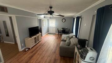 A living room with a television, a ceiling fan, a dining area with a table and chairs, and a kitchen area with a coffee maker.