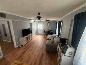 Ein Wohnzimmer mit Fernseher, Deckenventilator, Essbereich mit Tisch und Stühlen sowie Küchenzeile mit Kaffeemaschine.