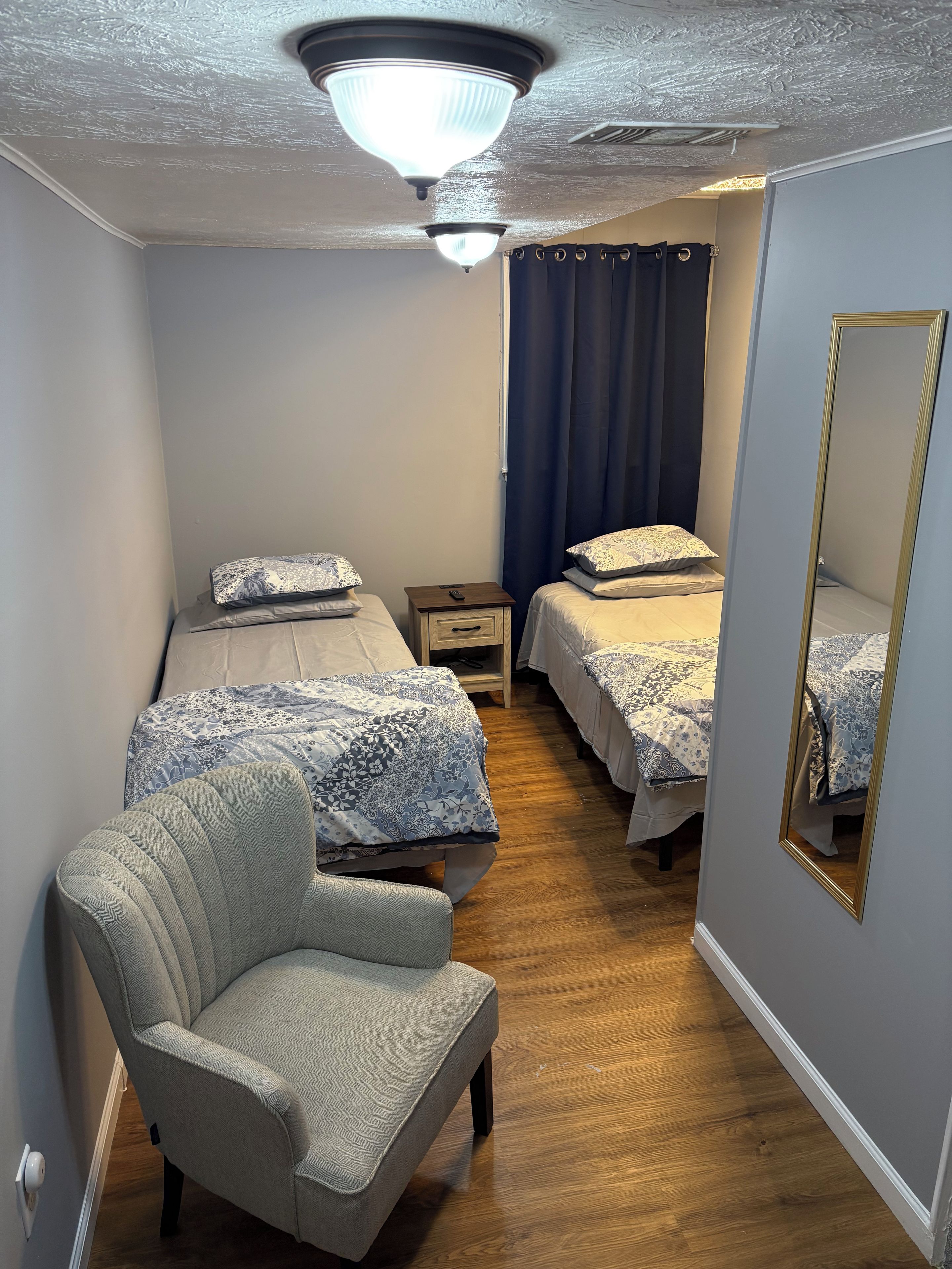 A bedroom with two beds, a chair, a nightstand, a mirror, and a ceiling light.