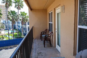 A balcony with two chairs and a table, overlooking a pool and a building with balconies. - Affordable 2-BR Escape at Surfside 1 #310 - 2 Minutes from the Sand (South Padre Island)