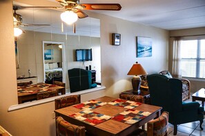 A room with a dining table, chairs, a mirror, a ceiling fan, a television, and artwork on the wall. - Affordable 2-BR Escape at Surfside 2 #107 - Steps from the Sand (South Padre Island)