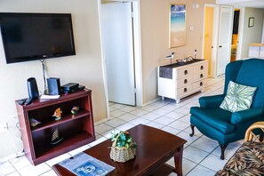 A living room with a TV, a wooden cabinet, a blue armchair, and a coffee table. - Affordable 2-BR Escape at Surfside 2 #107 - Steps from the Sand (South Padre Island)