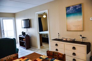 A room with a television, a desk with a lamp, a bed, a dresser, and a painting on the wall. - Affordable 2-BR Escape at Surfside 2 #107 - Steps from the Sand (South Padre Island)