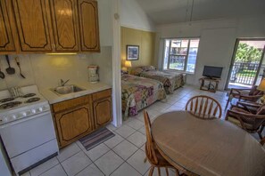 Een kleine keuken met een fornuis, gootsteen en houten kastjes, een eethoek met tafel en stoelen, en een slaapkamer met een bed en raam.