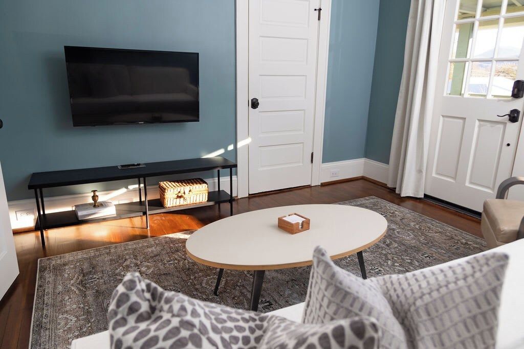 Een woonkamer met een flatscreen televisie aan de muur, een ronde salontafel en een bank met gekleurde kussens.