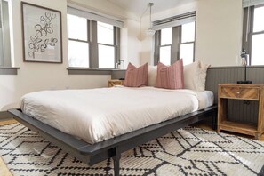 A bed with a headboard, a bedside table, and a wall-mounted lamp. - Stay Roanoke | City Loft w/ Deck & Free Parking (Roanoke)