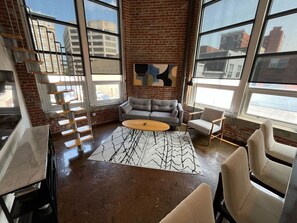 A spacious room with a brick wall, large windows, a gray sofa, a round coffee table, and a patterned rug. - Stay Roanoke | Chic downtown 1BR with study loft (Roanoke)
