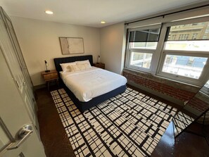 A bedroom with a bed, a nightstand, a window, and a patterned rug. - Stay Roanoke | Chic downtown 1BR with study loft (Roanoke)