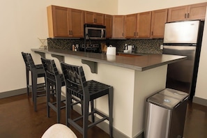A kitchen with wooden cabinets, a stainless steel refrigerator, a microwave, and a bar stool area. - Stay Roanoke | Downtown Loft, Fast WiFi & Views (Roanoke)