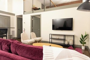 A living room with a large flat-screen TV mounted on the wall, a sofa, two chairs, a coffee table, and a plant. - Stay Roanoke | Downtown Loft, Fast WiFi & Views (Roanoke)
