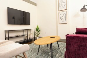 Ein Wohnzimmer mit einem wandmontierten Flachbildfernseher, einem runden Holztisch, einem roten Samtsofa und einem schwarzen Metallkonsole mit Dekorationselementen.