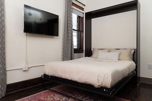 A bedroom with a bed, a television mounted on the wall, a window with curtains, and a patterned rug. - Stay Roanoke | Stylish historic loft near downtown (Roanoke)