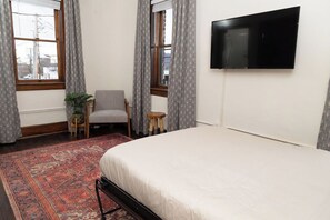 A bedroom with a bed, a TV mounted on the wall, a chair, a small table, and a window with curtains. - Stay Roanoke | Stylish historic loft near downtown (Roanoke)