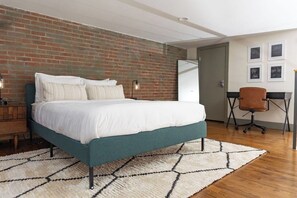A bedroom with a brick wall, a bed with white bedding, a desk with a chair, and a white rug. - Stay Roanoke | Downtown Loft, Fast WiFi, City View (Roanoke)