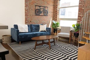 A living room with a blue sofa, a wooden coffee table, a striped rug, and a brick wall with a bicycle decoration. - Stay Roanoke | Downtown Loft, Fast WiFi, City View (Roanoke)