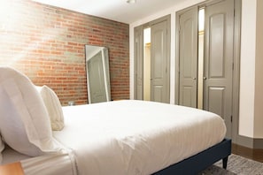 A bedroom with a brick wall, a bed with white bedding, a mirror, and a set of grey doors. - Stay Roanoke | Downtown Loft, Fast WiFi, City View (Roanoke)