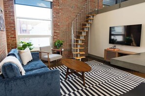 A modern living room with a blue sofa, a wooden coffee table, a black and white striped rug, a TV, and a brick wall. - Stay Roanoke | Downtown Loft, Fast WiFi, City View (Roanoke)