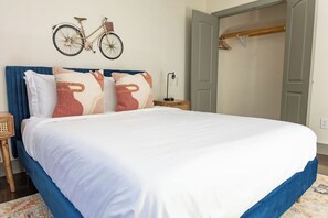 Ein ordentlich bezogenes Bett mit weißer Bettwäsche und Zierkissen, ein Nachttisch mit Lampe sowie ein an der Wand befestigtes Fahrrad.