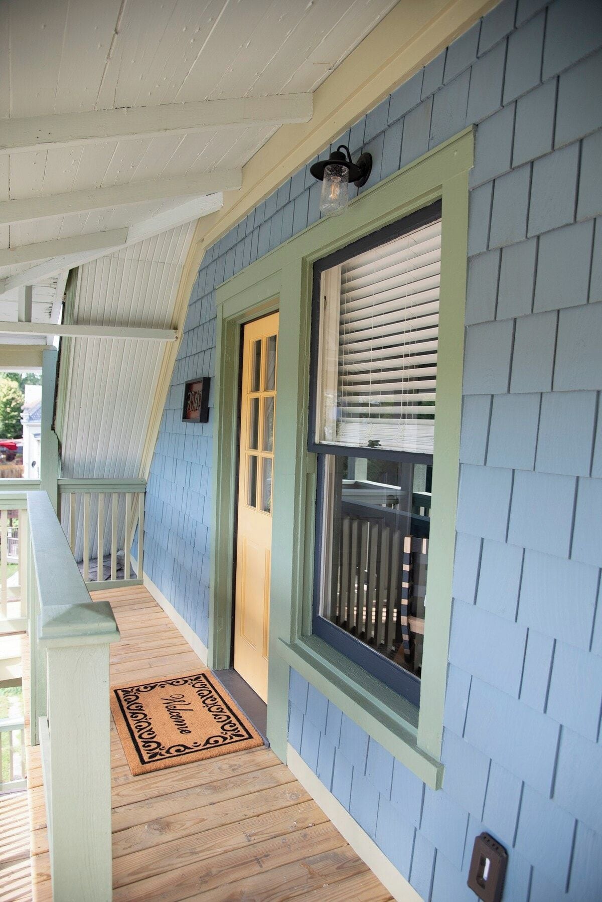A porch with a wooden floor, a welcome mat, and a window with blinds.